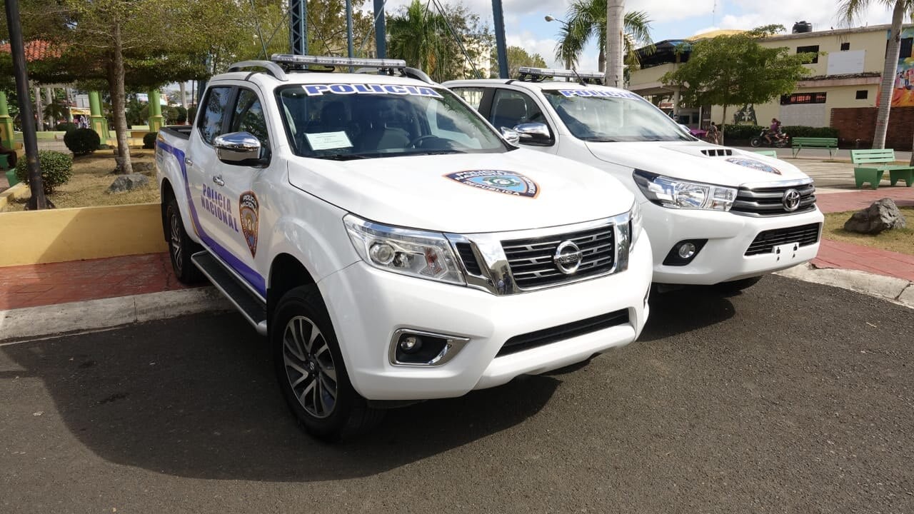 Two white Policía Nacional pickup trucks, a Nissan and a Toyota, with light bars, parked on asphalt beside a town square with palm trees
