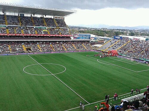 A packed grandstand at Estadio Ricardo Saprissa in Costa Rica, with fans filling the stands above a green pitch during a match.