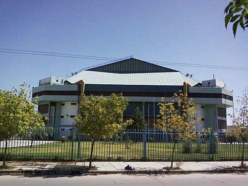 Exterior view of the Ruca Che indoor arena in Neuquén, Argentina, a large blue-roofed stadium behind a fence and trees.