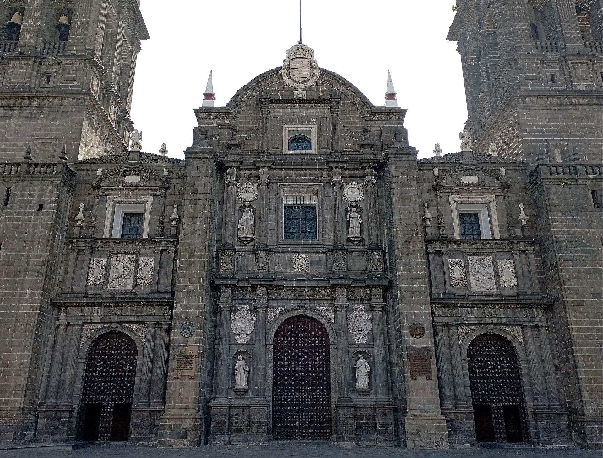 Grey stone facade of Puebla Cathedral with three arched, studded wooden doors, statues in niches and the base of two bell towers under a pale sky