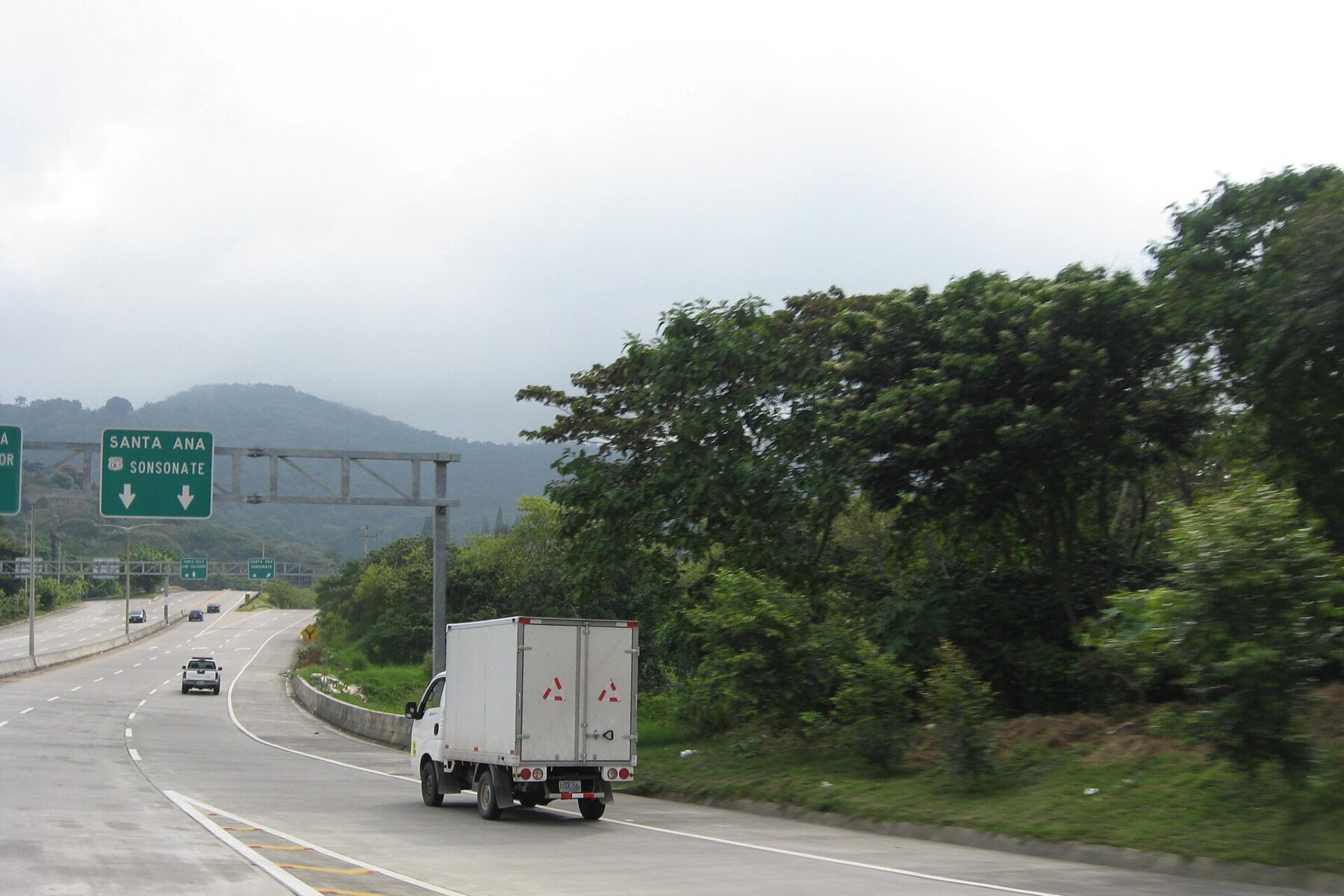 White box truck and cars on a multi-lane concrete road below a green overhead sign to Santa Ana and Sonsonate, wooded hills and low cloud behind