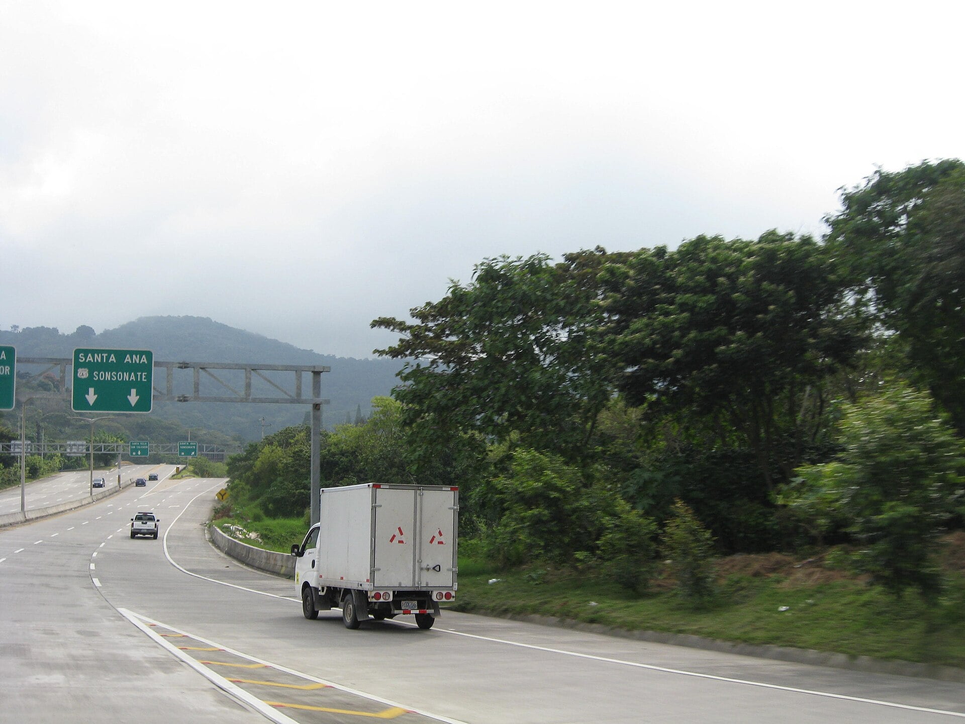 White box truck and cars on a multi-lane concrete road below a green overhead sign to Santa Ana and Sonsonate, wooded hills and low cloud behind