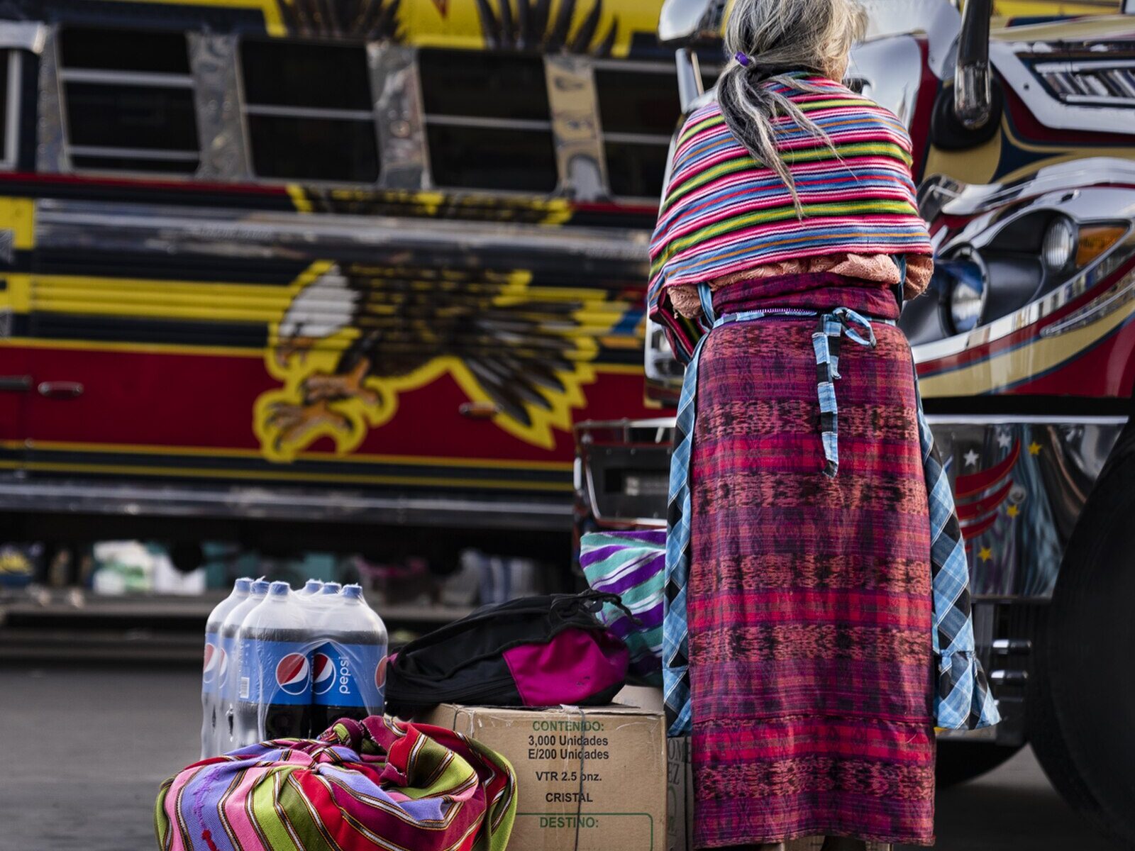 A woman with bundles waits beside painted buses in highland Guatemala