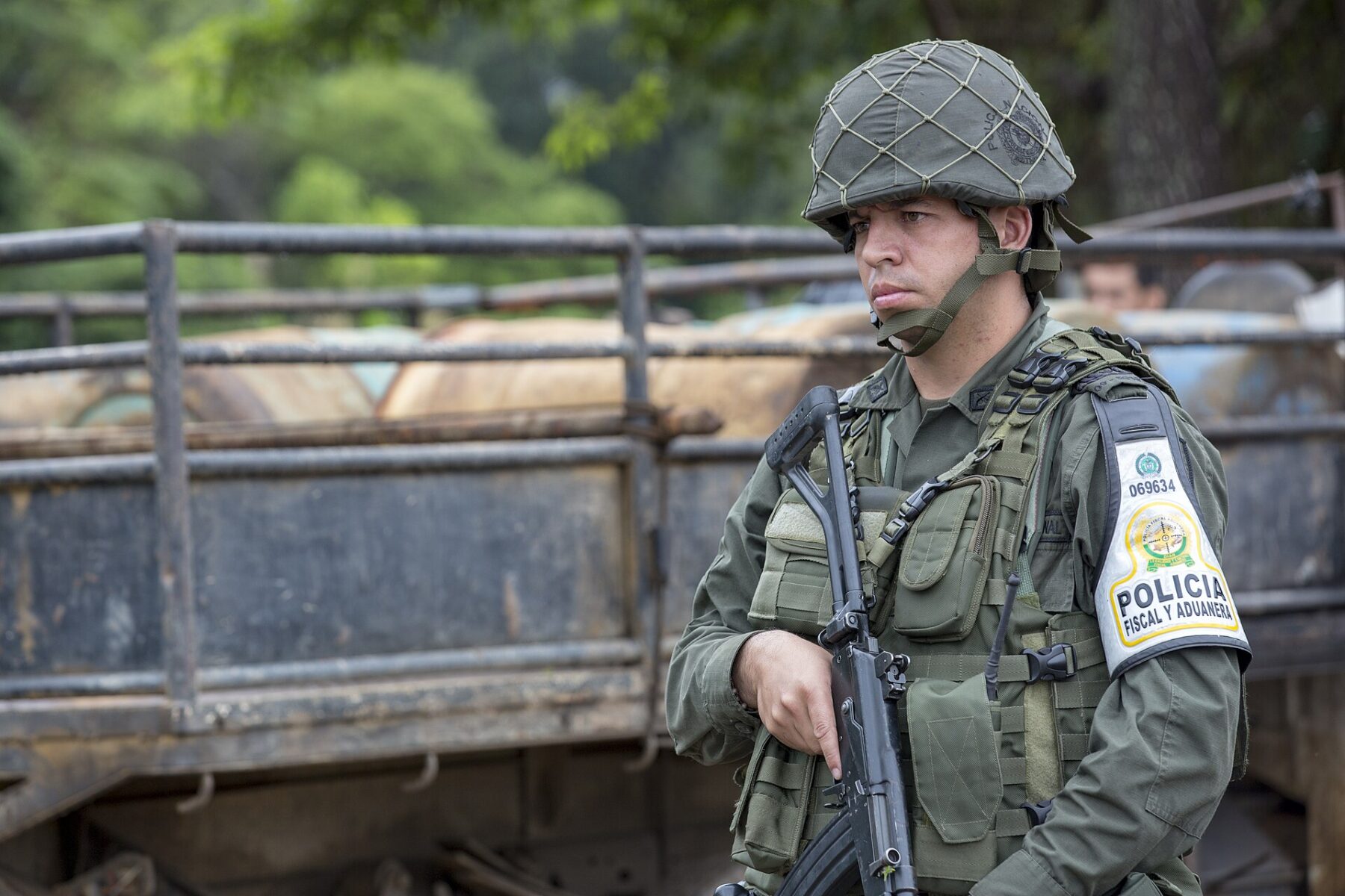 A Colombian national police officer in helmet and body armour holding a rifle, standing beside a truck
