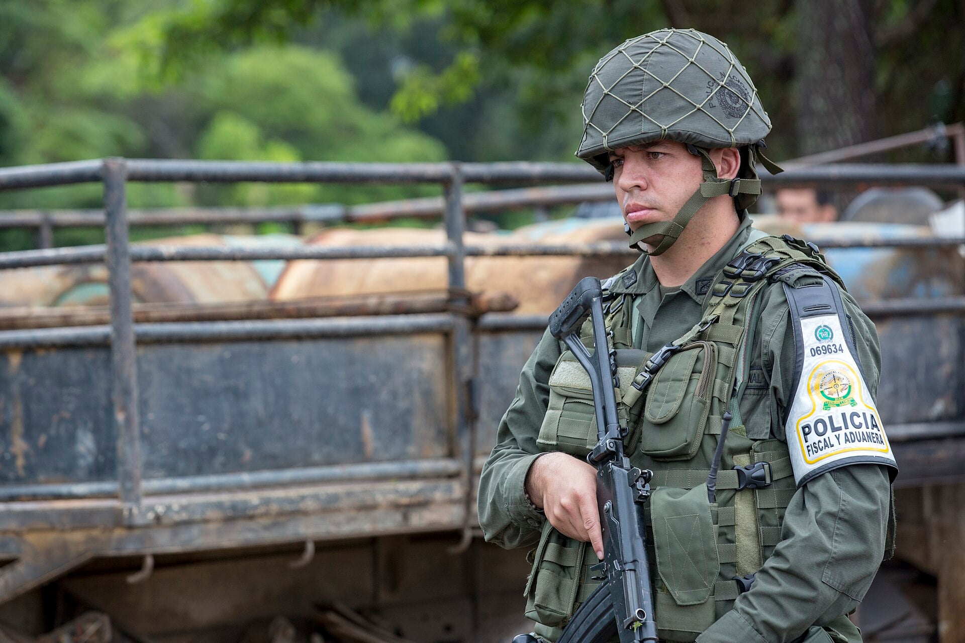 A Colombian national police officer in helmet and body armour holding a rifle, standing beside a truck