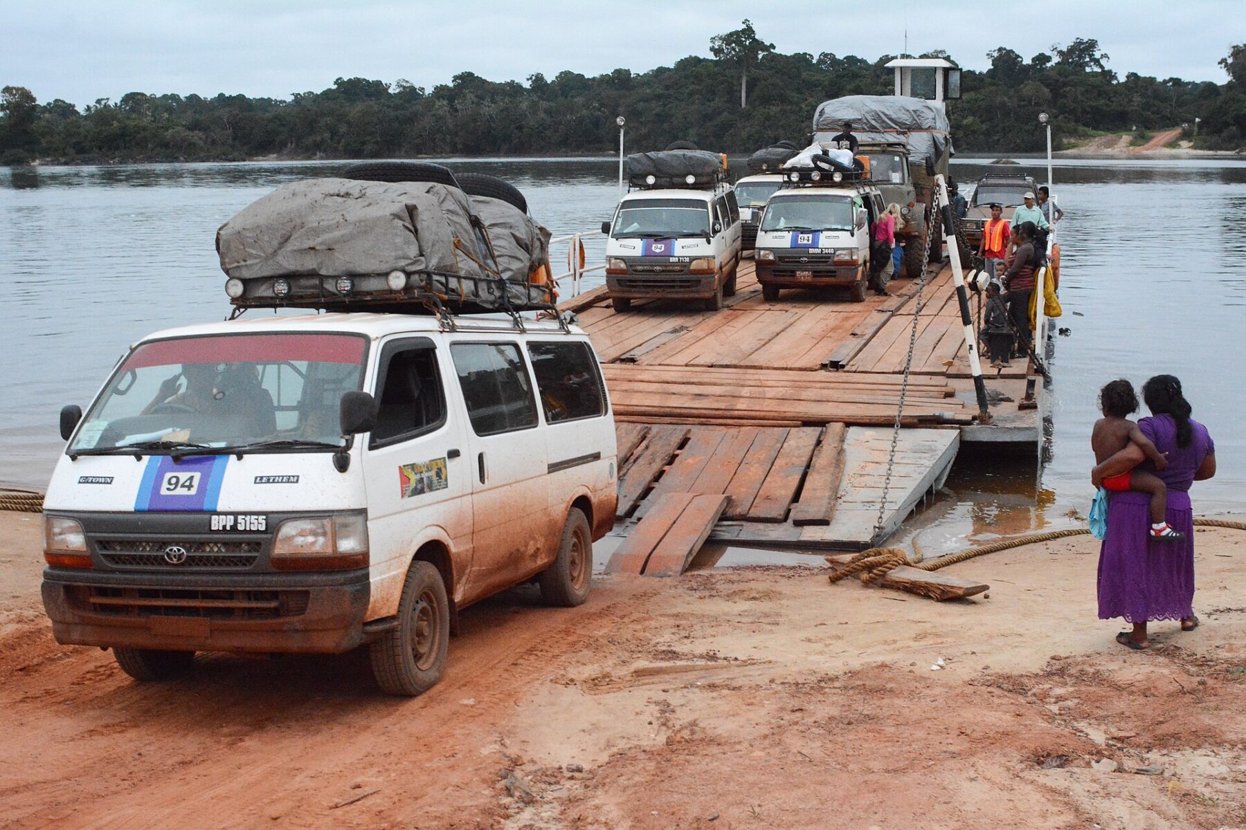 Minibuses driving off a wooden pontoon ferry onto a red dirt riverbank in the Guyanese interior