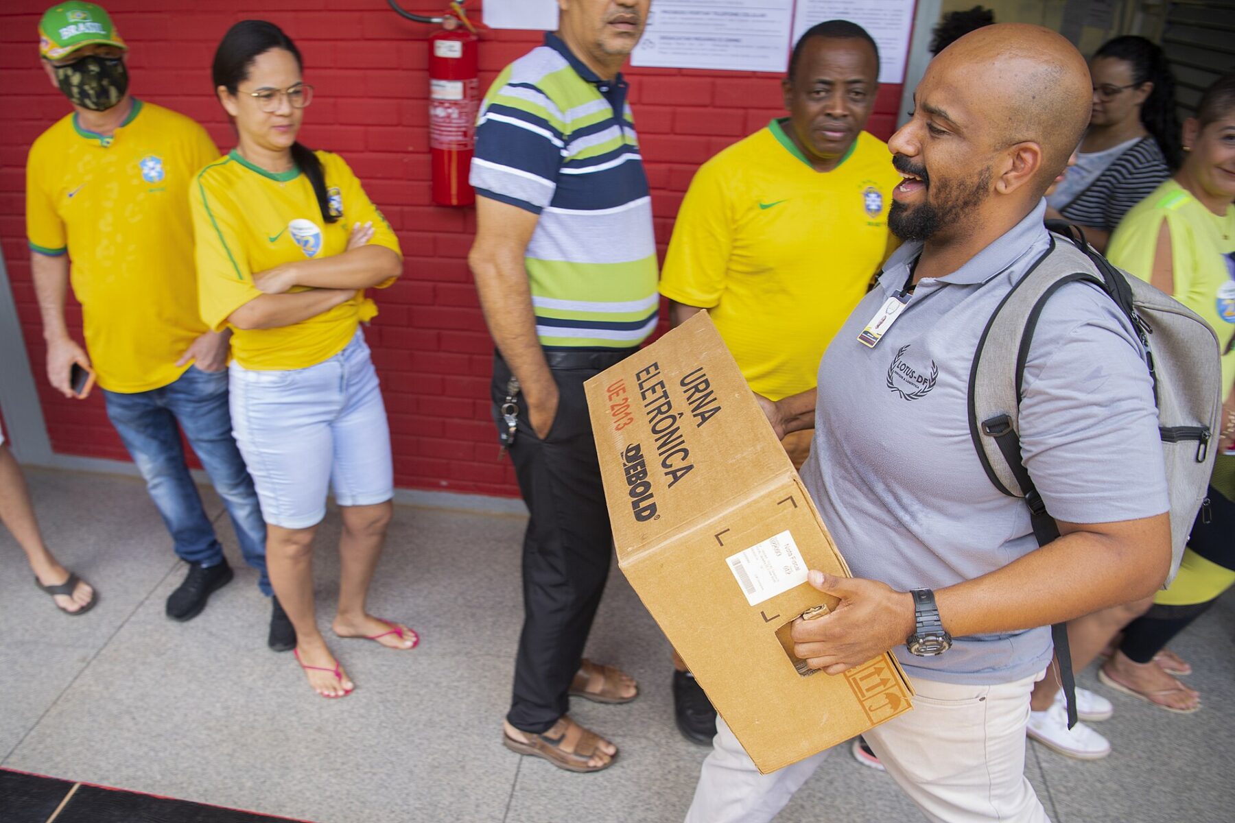 Voters in Brazil football shirts queue outside a polling station as an official carries a boxed electronic voting machine