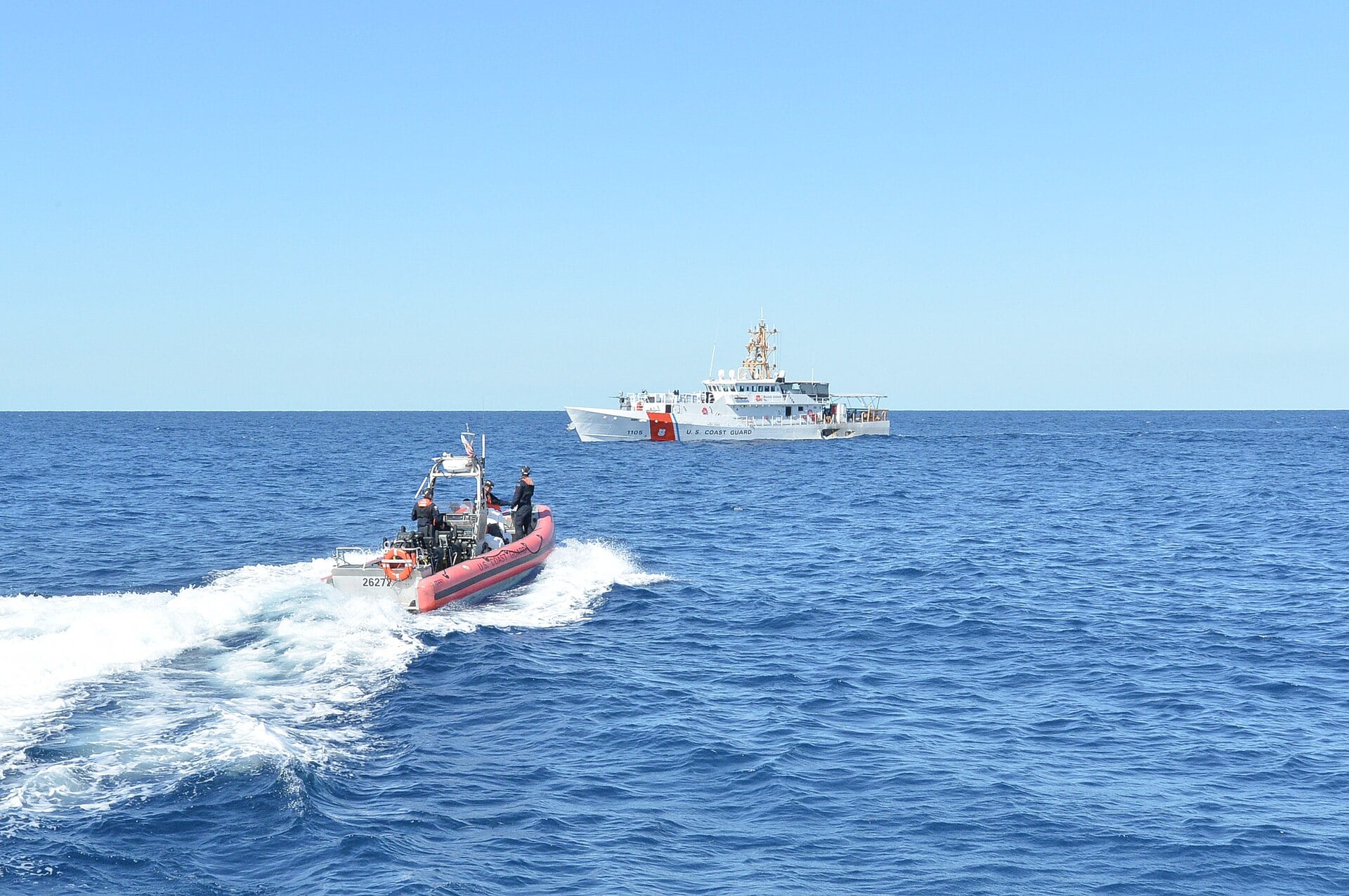 A US Coast Guard cutter at sea on calm blue water, with a small orange-sided Coast Guard boat speeding toward it