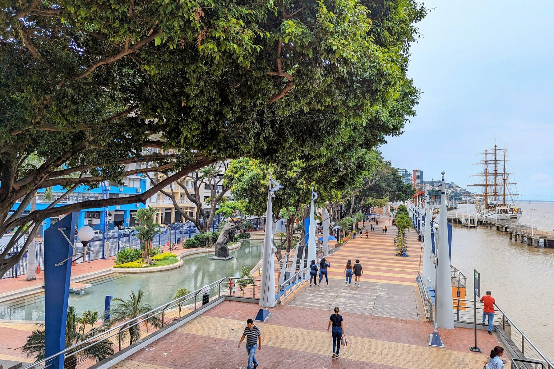 Waterfront promenade with people, trees, and a tall ship