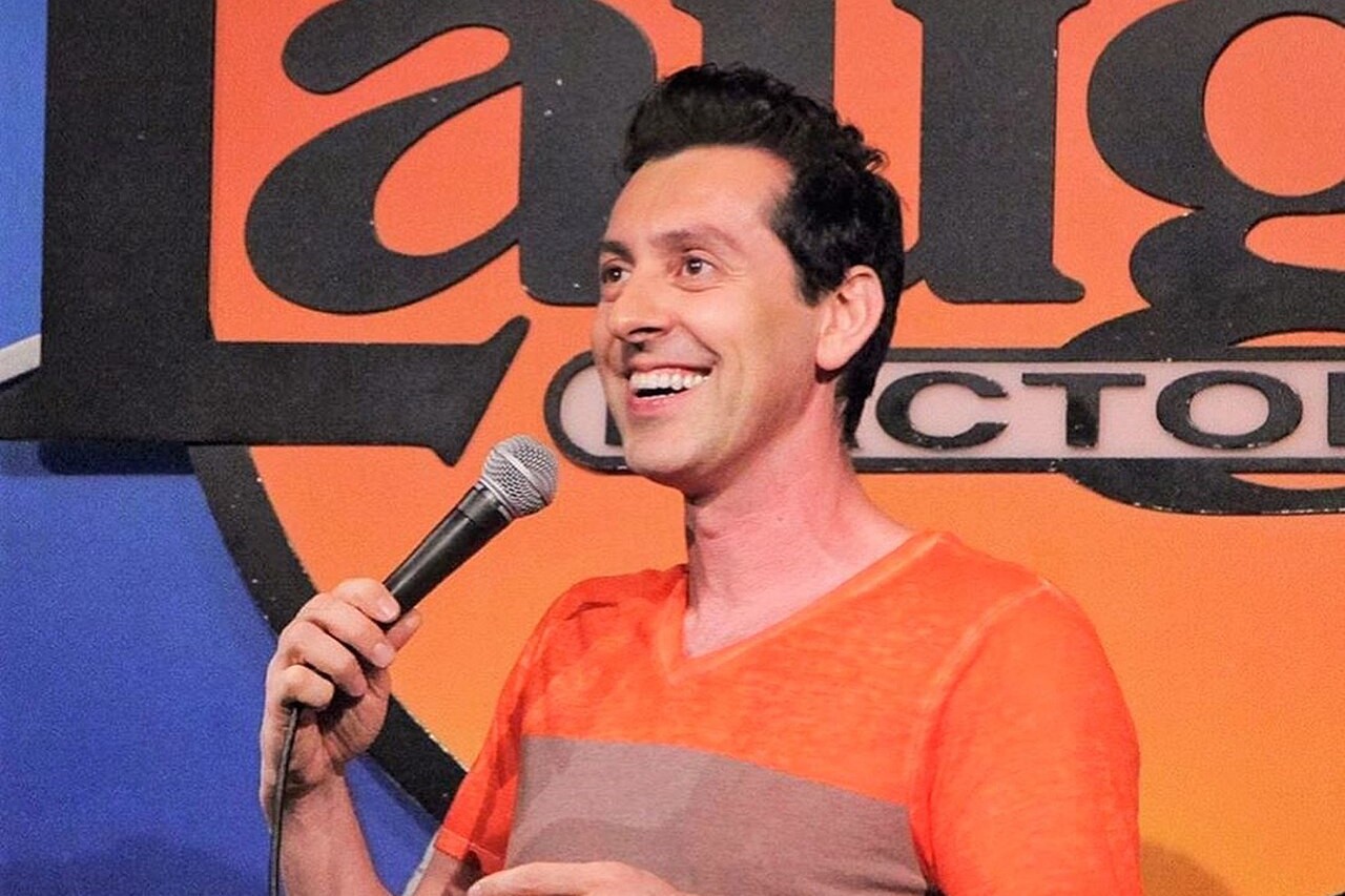 Comedian Max Amini smiling on stage with a microphone in front of a Laugh Factory sign