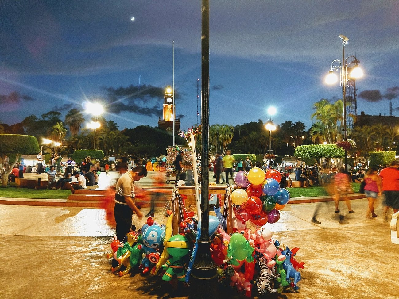 Crowds and a balloon seller on the Plaza Grande in Mérida, Yucatán, at night, with a lit church tower behind.