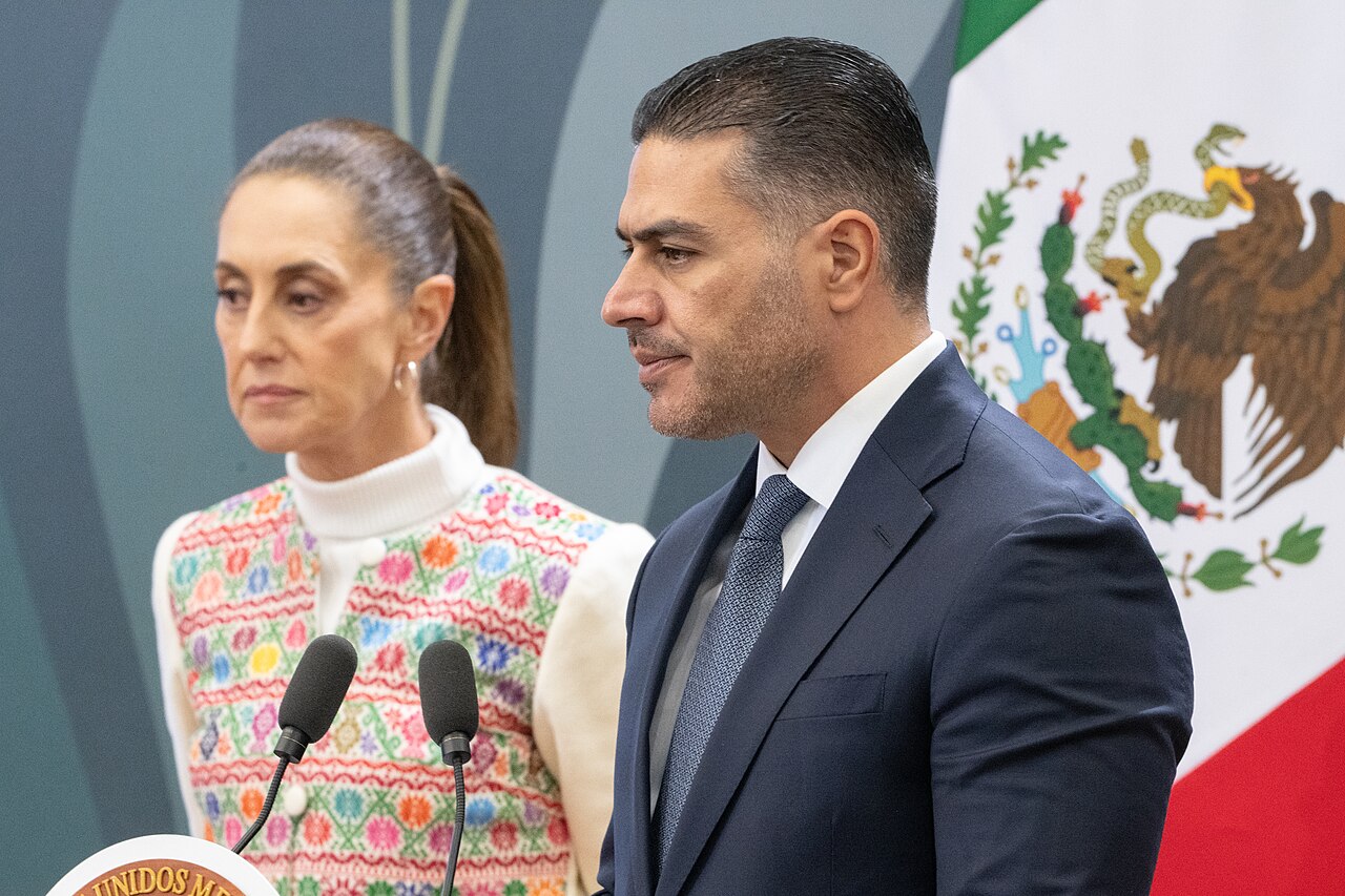 Mexico homicides briefing: Security Minister Omar García Harfuch speaks at a podium beside President Claudia Sheinbaum, with the Mexican flag behind them