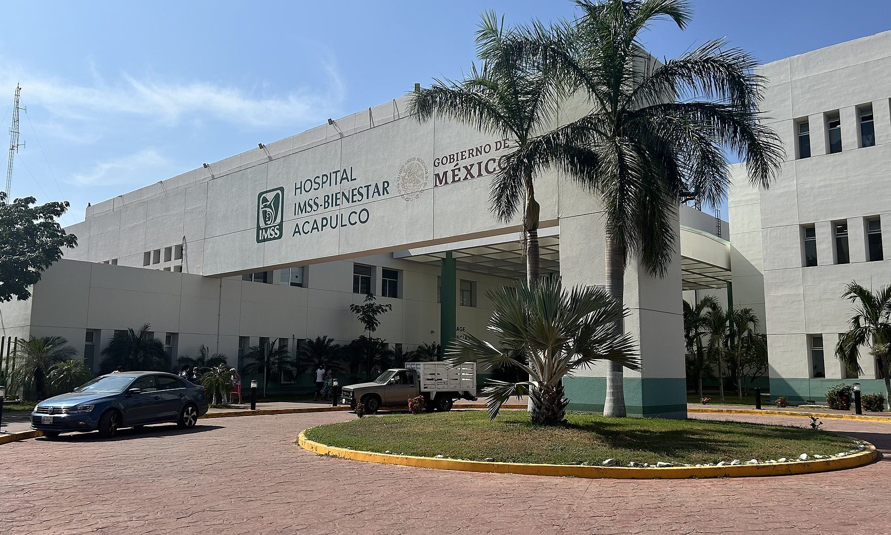 White front of the IMSS-Bienestar hospital in Acapulco, Mexico, with palm trees, a roundabout and parked cars