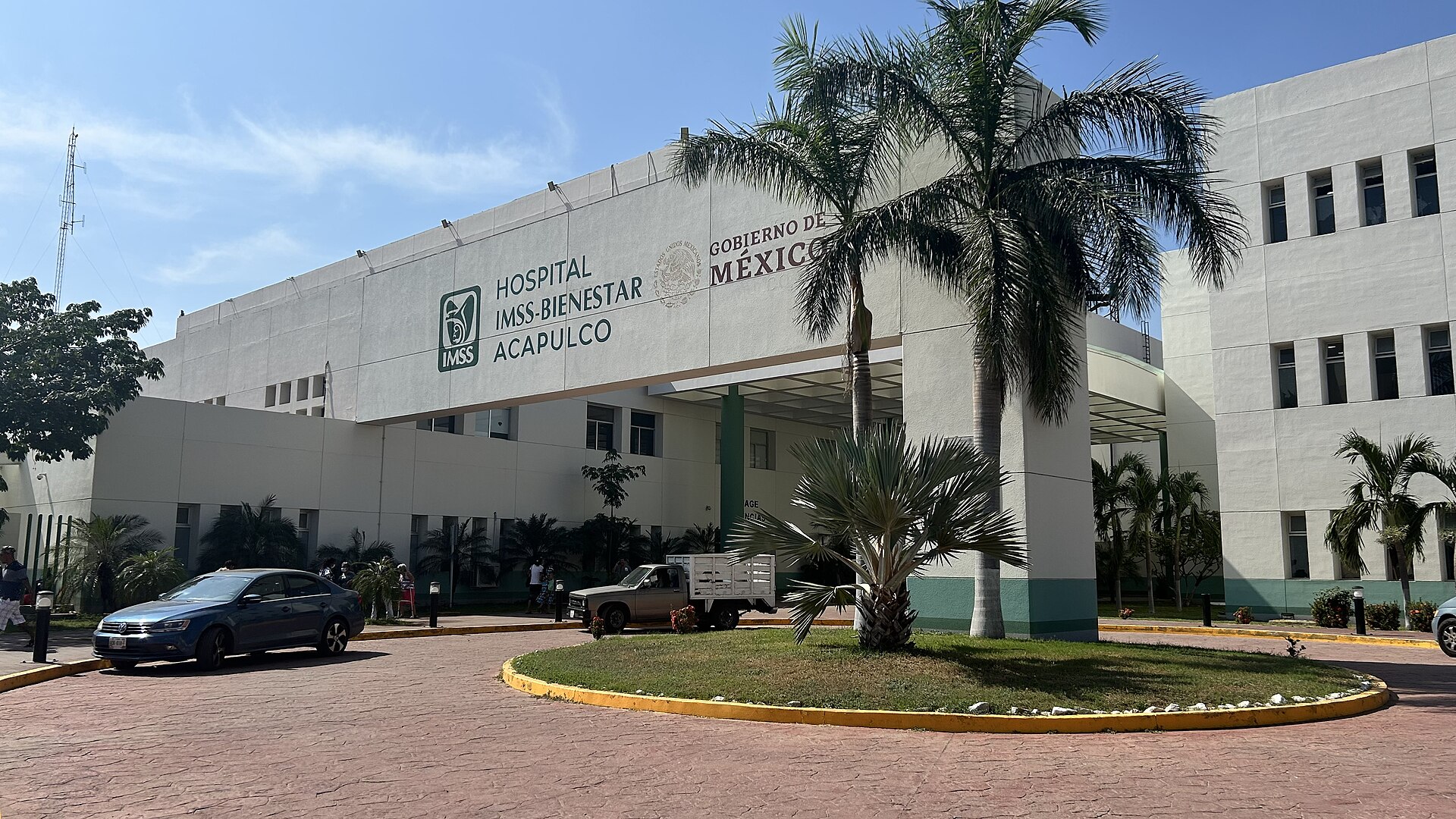 White front of the IMSS-Bienestar hospital in Acapulco, Mexico, with palm trees, a roundabout and parked cars