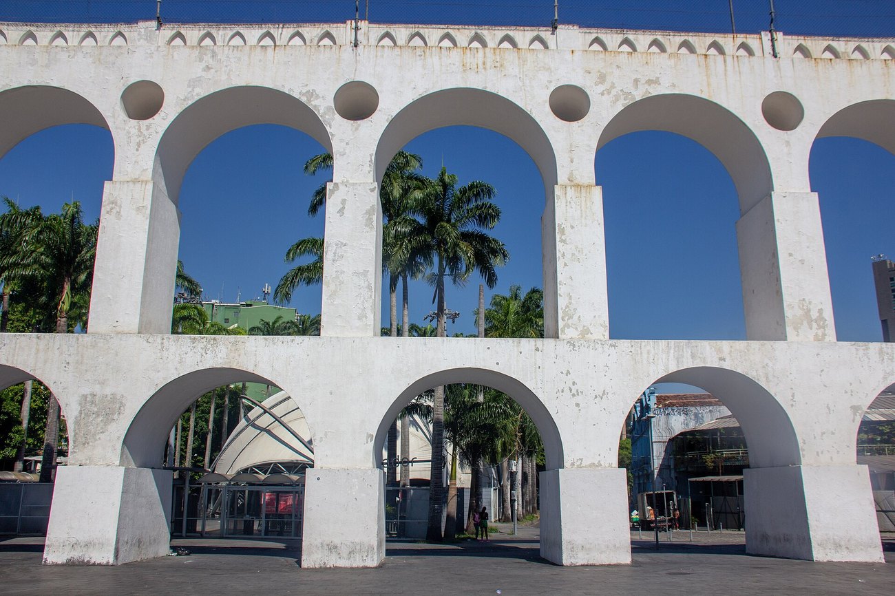The white arches of the Arcos da Lapa in Rio de Janeiro, with palm trees and the tent roof of the Circo Voador behind them.