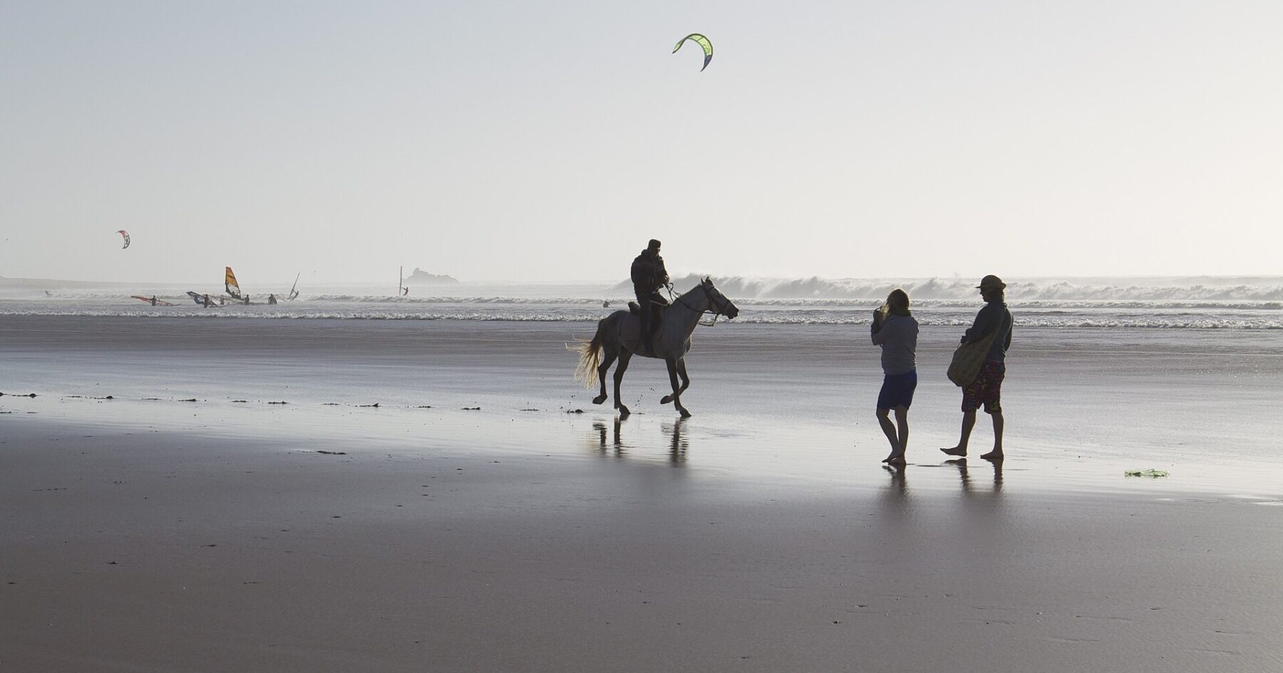 A horse rider and two walkers on Essaouira beach, Morocco, with kitesurfers offshore, near the site of MOGA Festival 2026