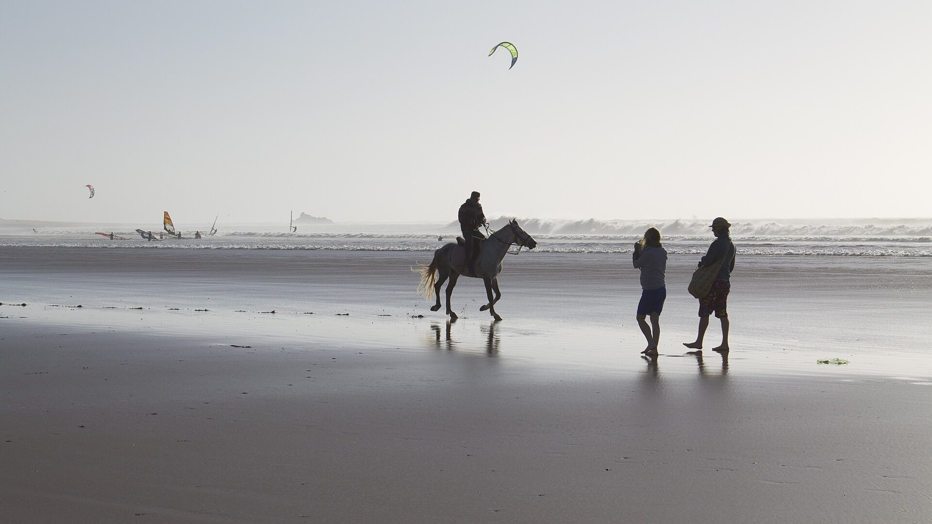 A horse rider and two walkers on Essaouira beach, Morocco, with kitesurfers offshore, near the site of MOGA Festival 2026