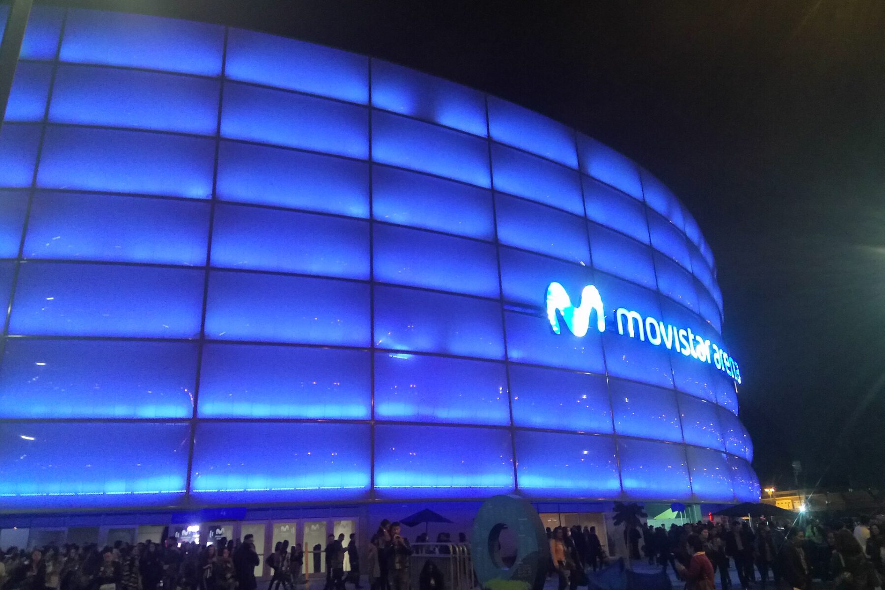 Movistar Arena in Bogotá lit blue at night, with its logo on the facade and a crowd queuing outside the entrance.