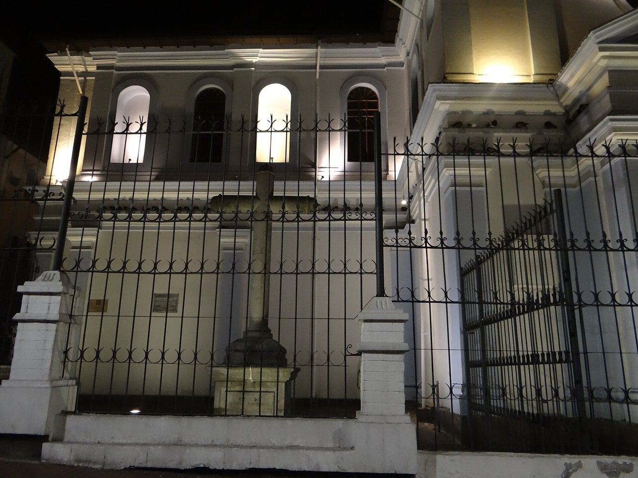A colonial church facade lit at night behind iron railings in the historic centre of Quito, with a stone cross in the forecourt.