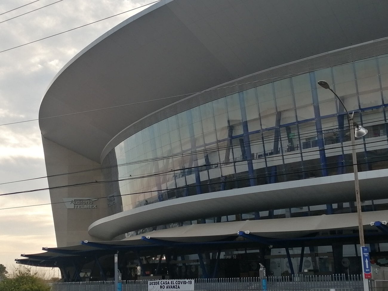The curved glass facade and cantilevered roof of the Auditorio Telmex in Zapopan, Guadalajara, seen from the street.