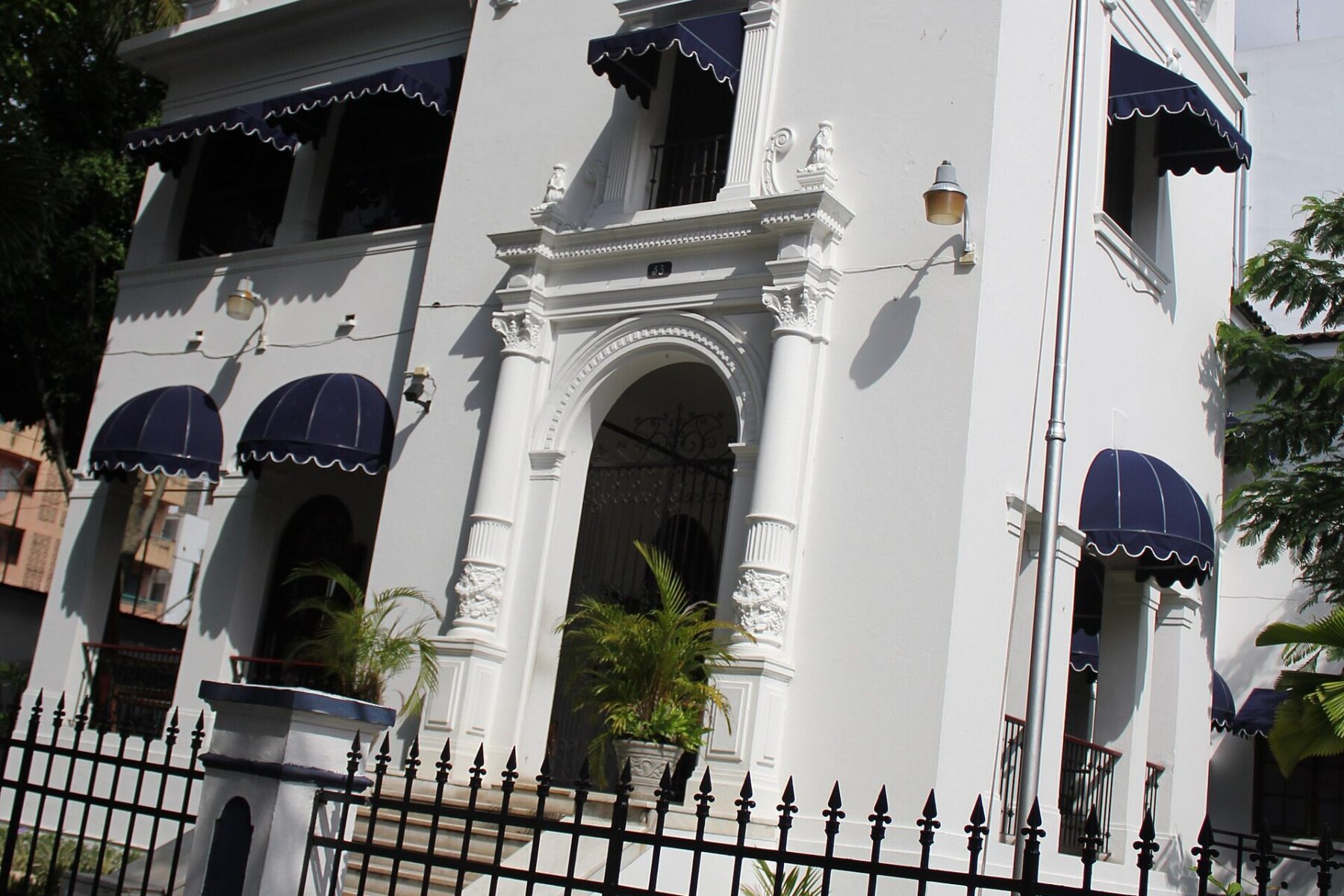 White colonial-style building with navy-blue scalloped awnings, an arched entrance between carved columns and a black iron fence, seen at an angle