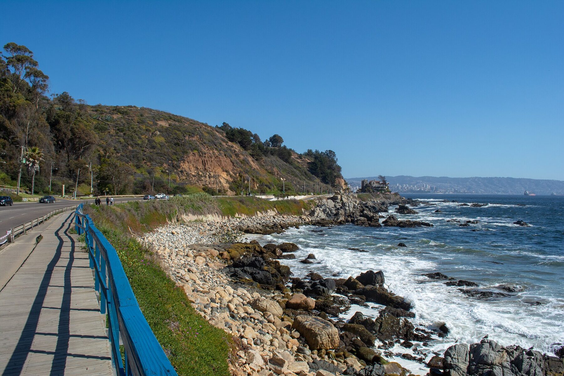 The coast road and wooden boardwalk at Reñaca, with a rocky shoreline, a green headland and Viña del Mar in the distance.