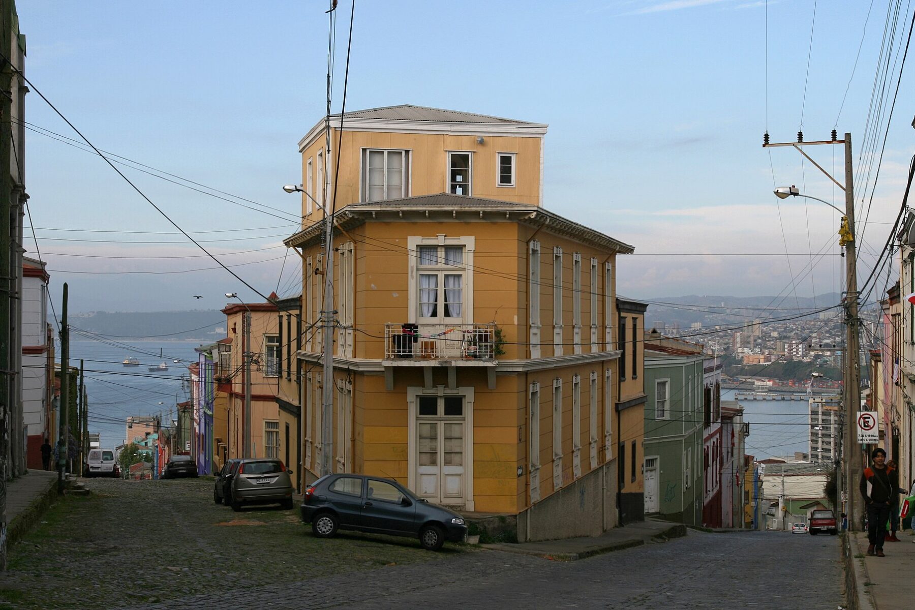 A cobbled street climbs a hillside in Valparaíso, Chile, with a yellow house at the corner.