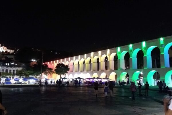 Rio de Janeiro nightlife: the Arcos da Lapa lit in pink, yellow and green at night, with people crossing the square and food stalls beneath the arches