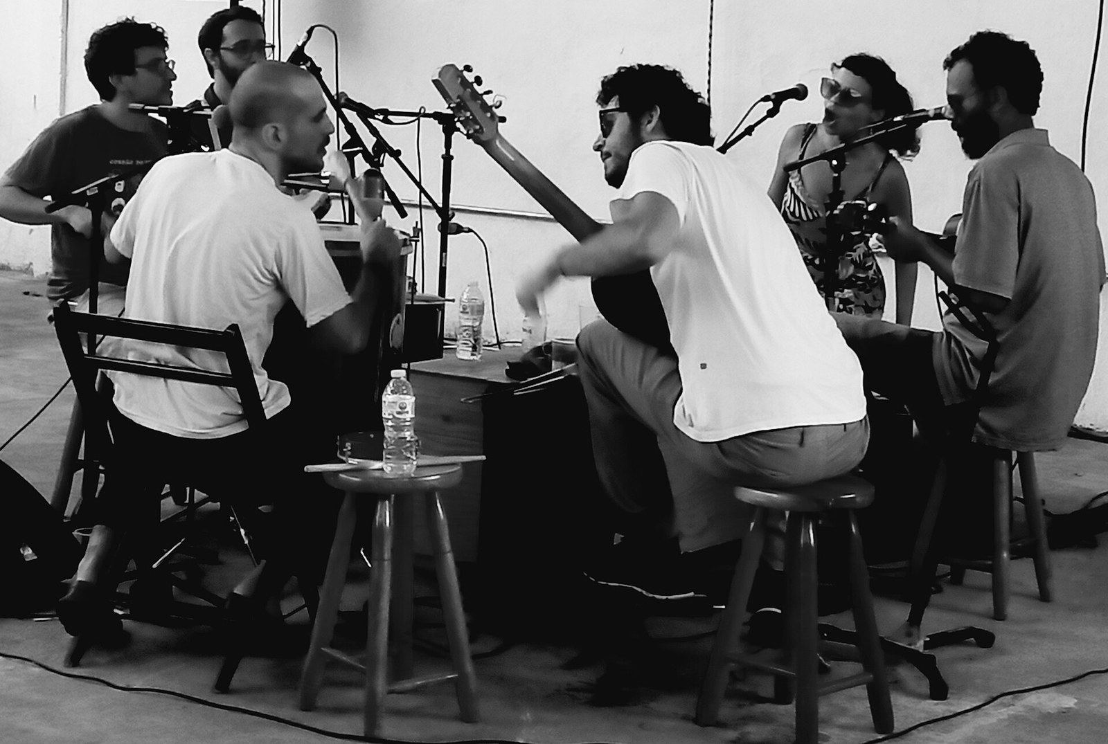 Black-and-white photo of samba musicians seated on stools around a table with guitars, percussion and microphones, a woman singing at the back