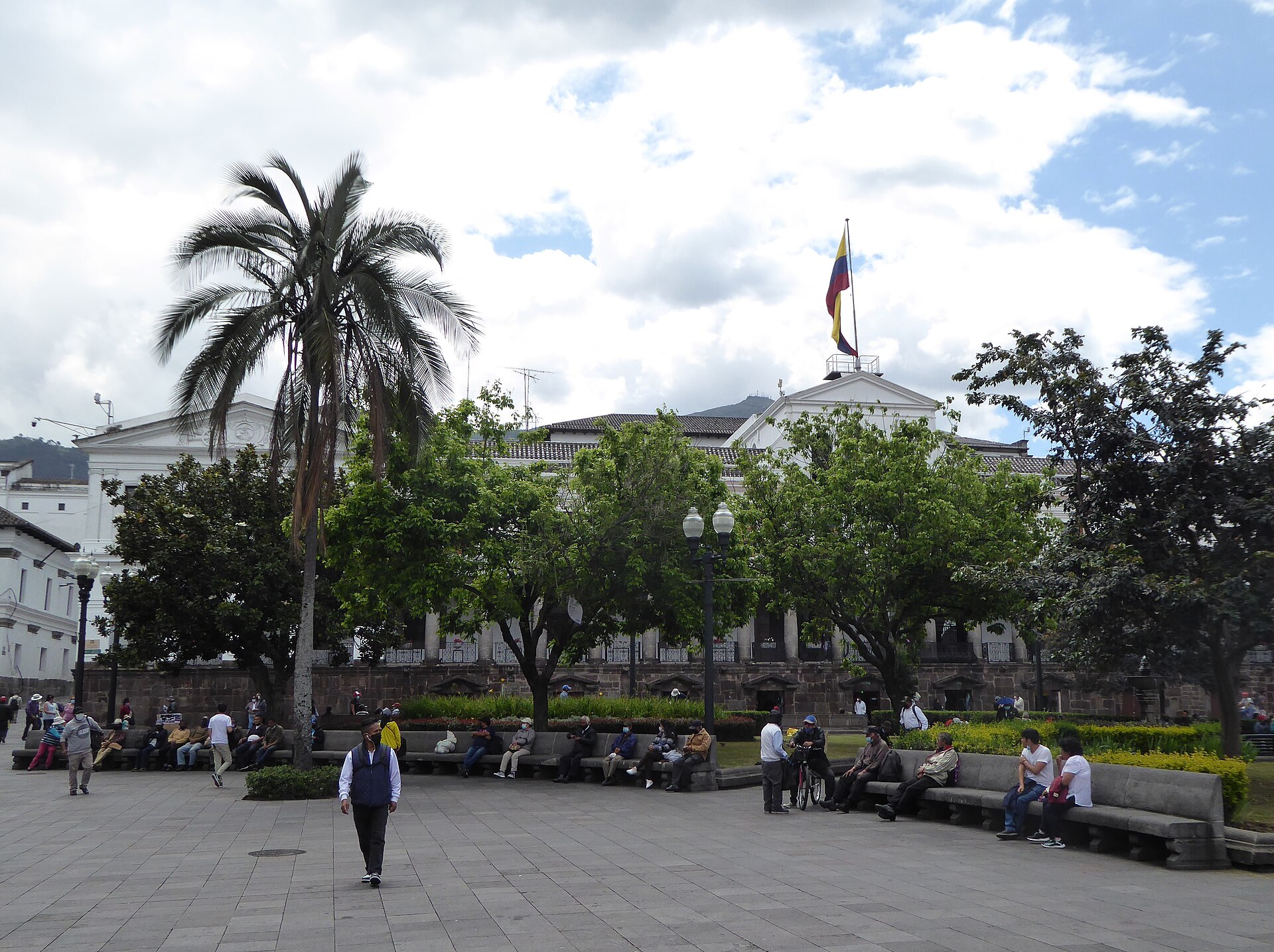People sitting on stone benches in Quito's Plaza Grande, with a palm tree and Carondelet Palace flying the Ecuadorian flag behind the trees