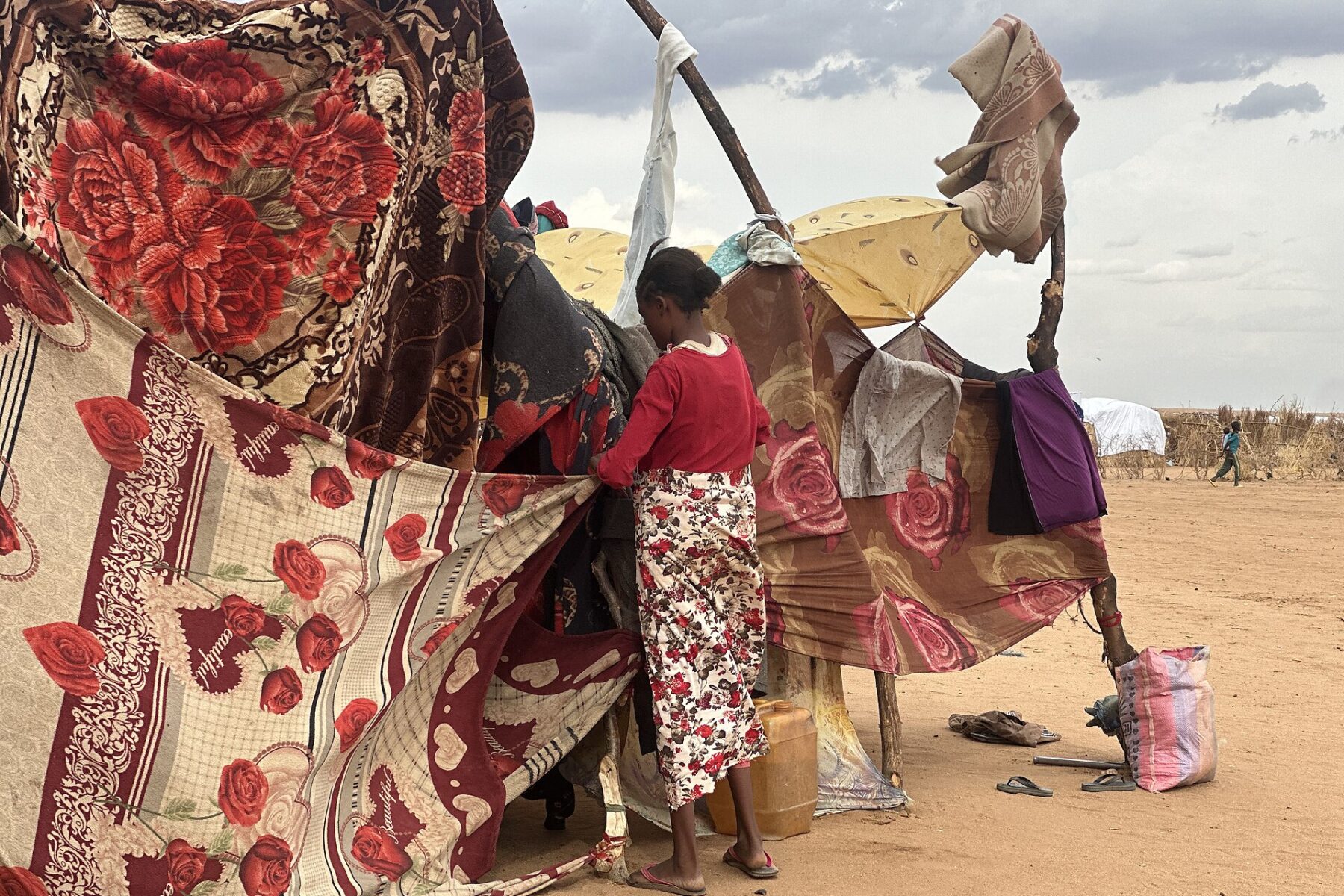 A displaced Sudanese woman stands beside a makeshift shelter made of pieces of cloth.