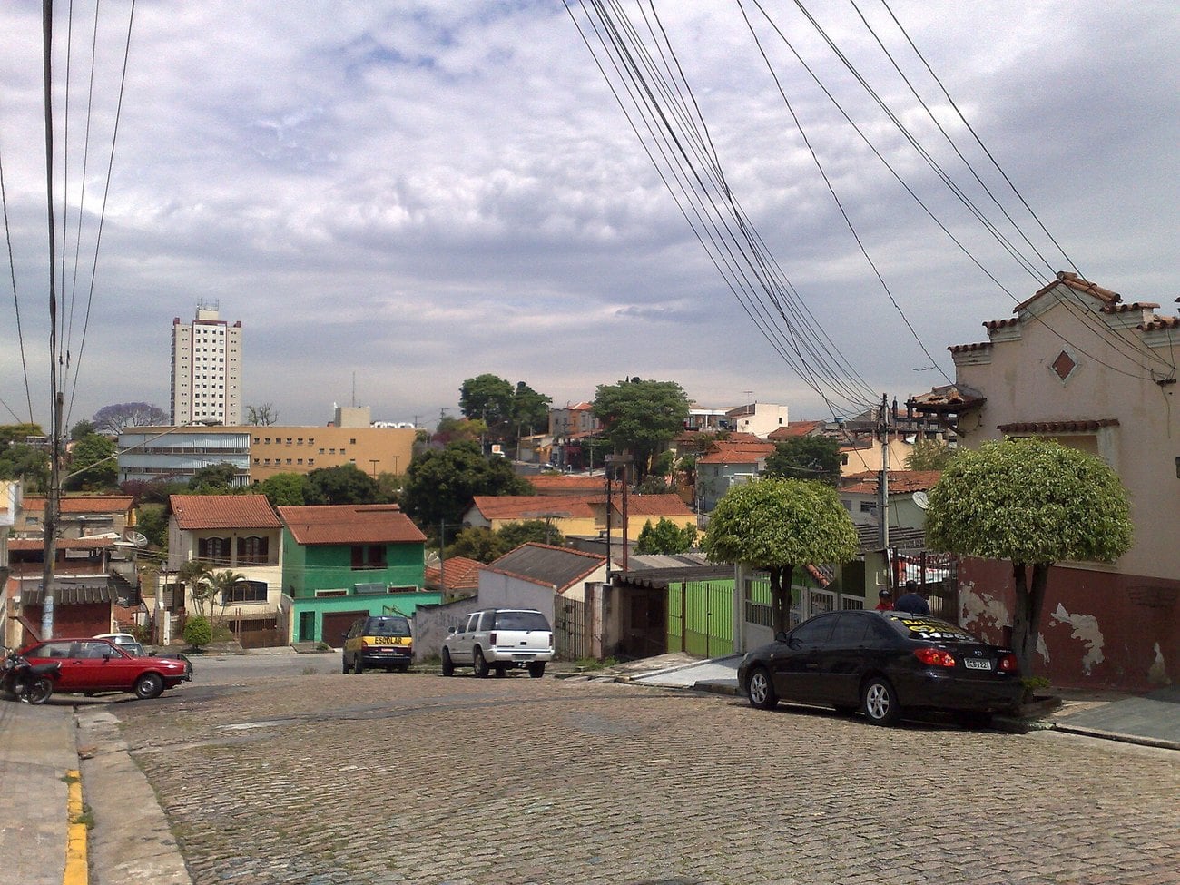 A cobbled residential street in the Penha district of Sao Paulo, with low houses, parked cars and overhead cables