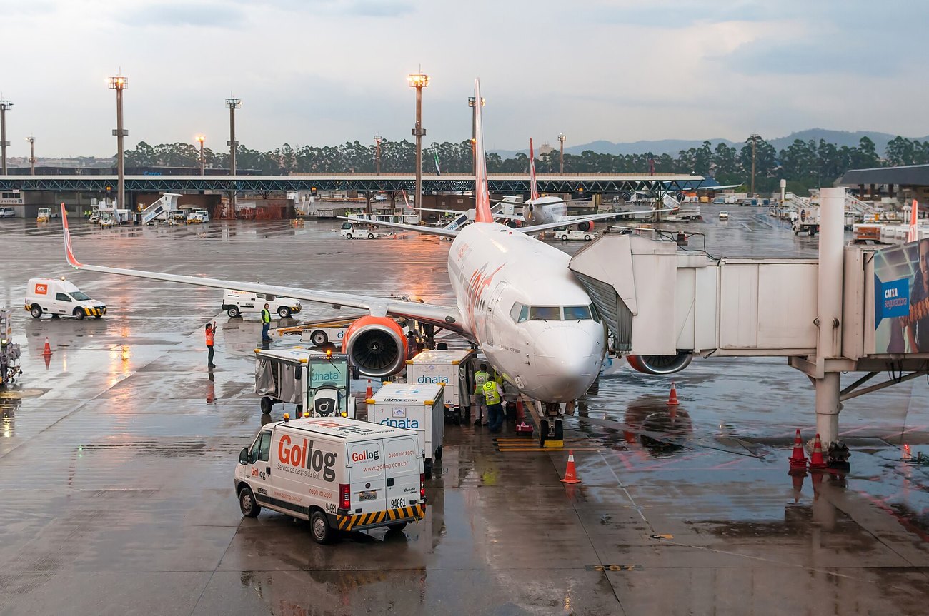 A GOL aircraft at a jet bridge on the wet apron at Guarulhos airport in Sao Paulo, with ground crew loading cargo at dusk.