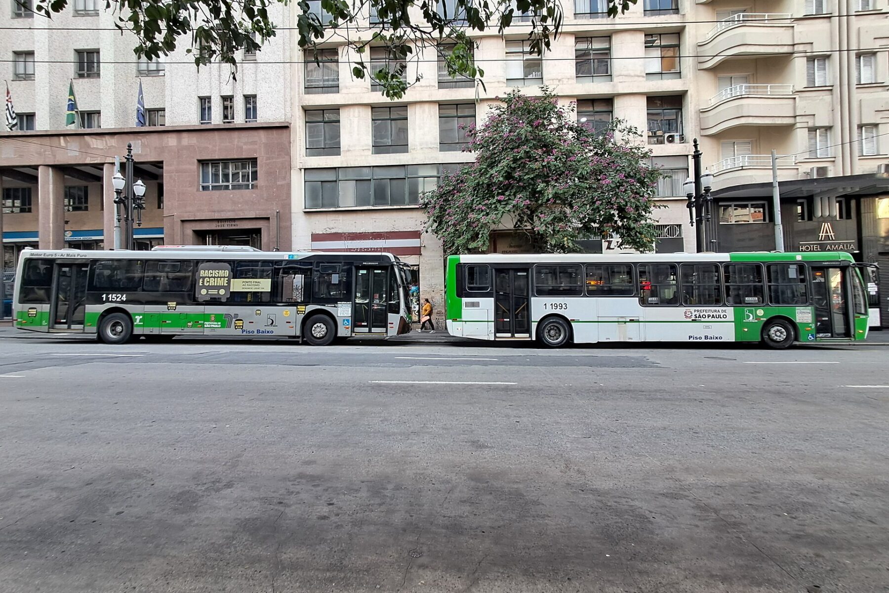 Two green and white São Paulo municipal buses stopped on a street in the city centre, with apartment blocks behind them.
