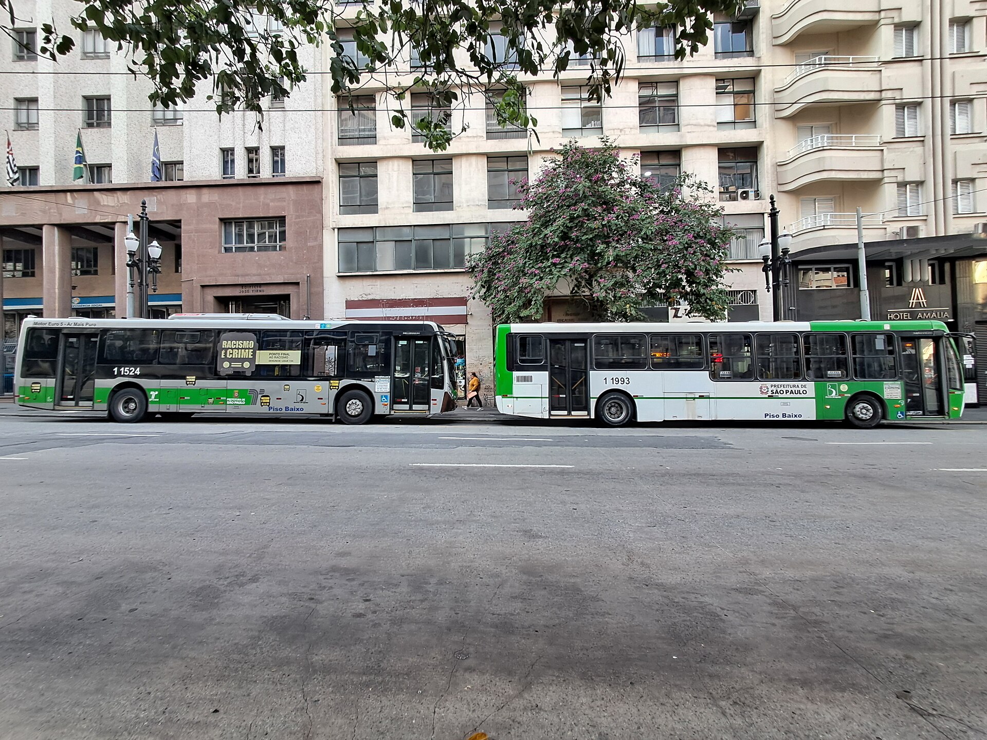Two green and white São Paulo municipal buses stopped on a street in the city centre, with apartment blocks behind them.