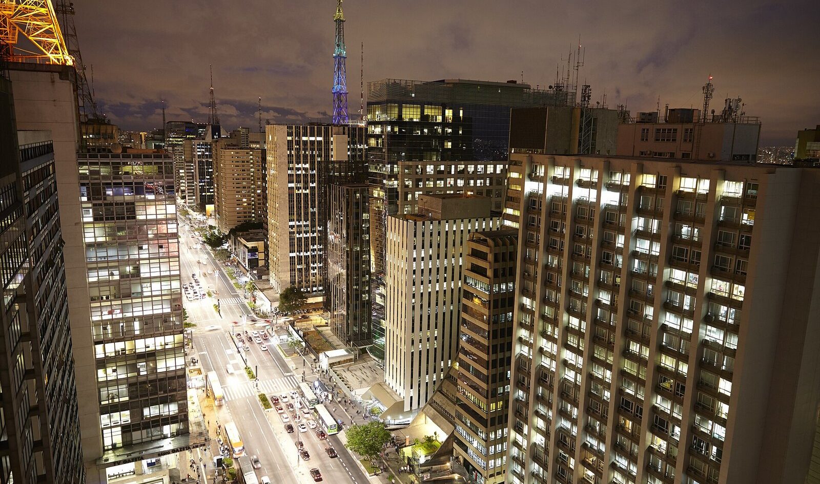 São Paulo nightlife: Avenida Paulista at night seen from above, with lit office towers, traffic and a floodlit broadcast mast