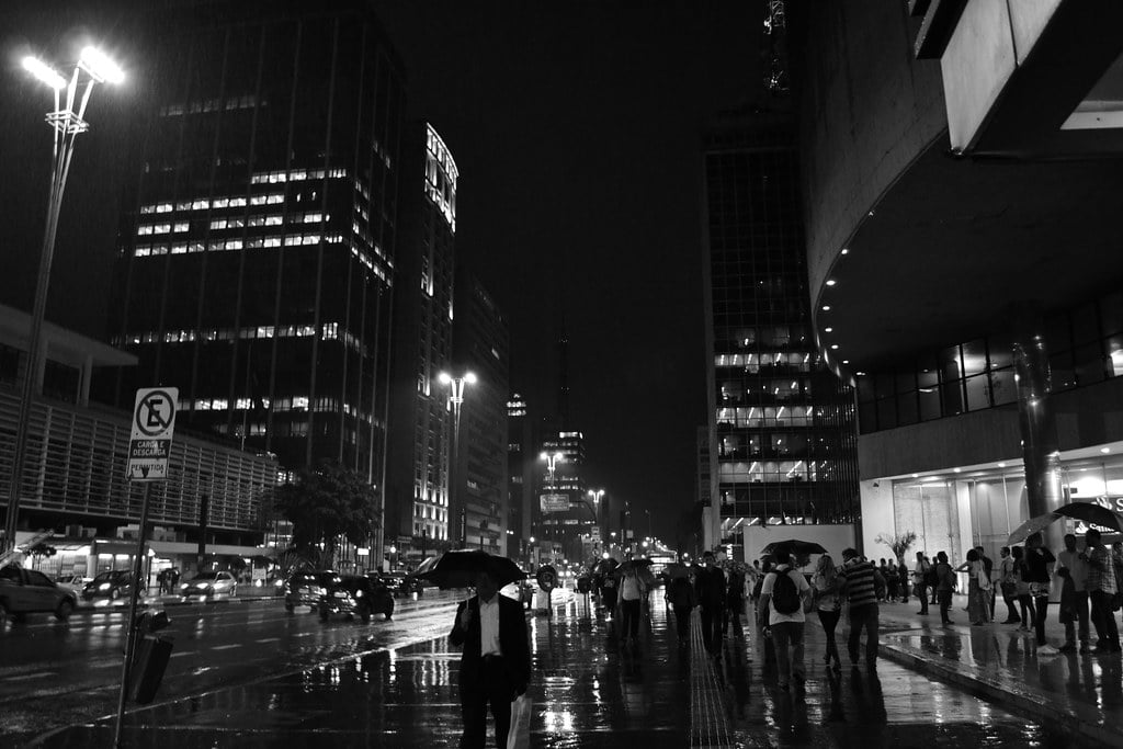 Avenida Paulista in São Paulo on a rainy night, with pedestrians under umbrellas and office towers lit up