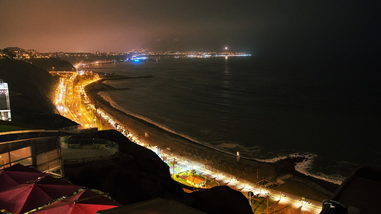 The lit coast road and beach below the cliffs of Miraflores in Lima at night, with the city glow behind