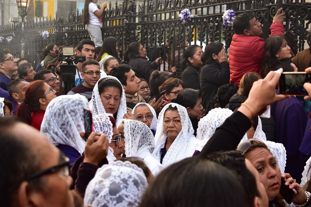 Women in white lace veils in the crowd at a Señor de los Milagros procession in Lima, with purple ribbons on the railings behind them.