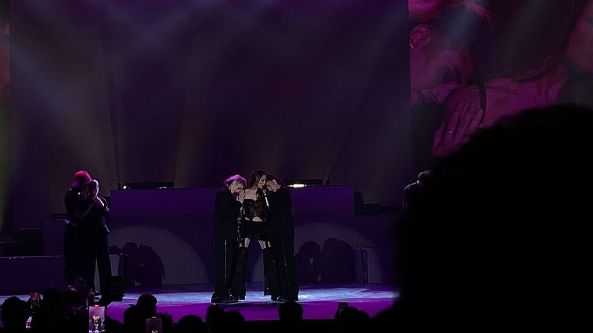 Tini on a purple-lit stage with two dancers, seen over the heads of the audience