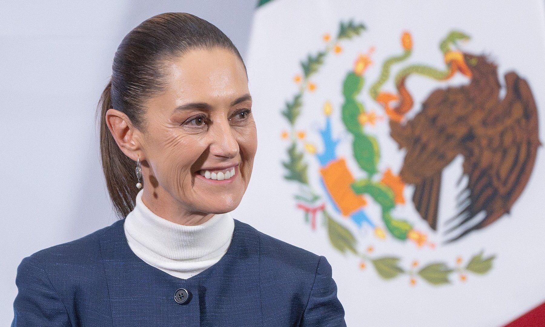 Mexican President Claudia Sheinbaum smiling in front of the Mexican flag and national coat of arms