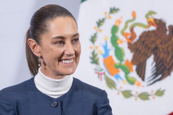 Mexican President Claudia Sheinbaum smiling in front of the Mexican flag and national coat of arms