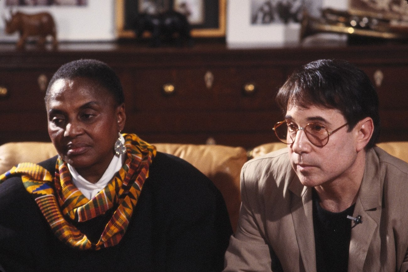 Miriam Makeba and Paul Simon sitting together on a sofa during an interview, Makeba wearing a kente scarf.