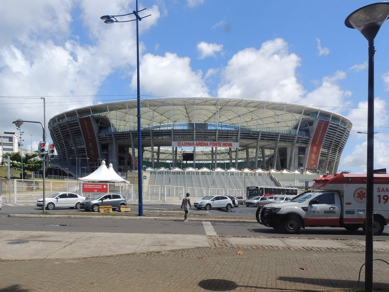 The Arena Fonte Nova in Salvador seen from the street, with its open roof ring, parked cars and an ambulance outside.
