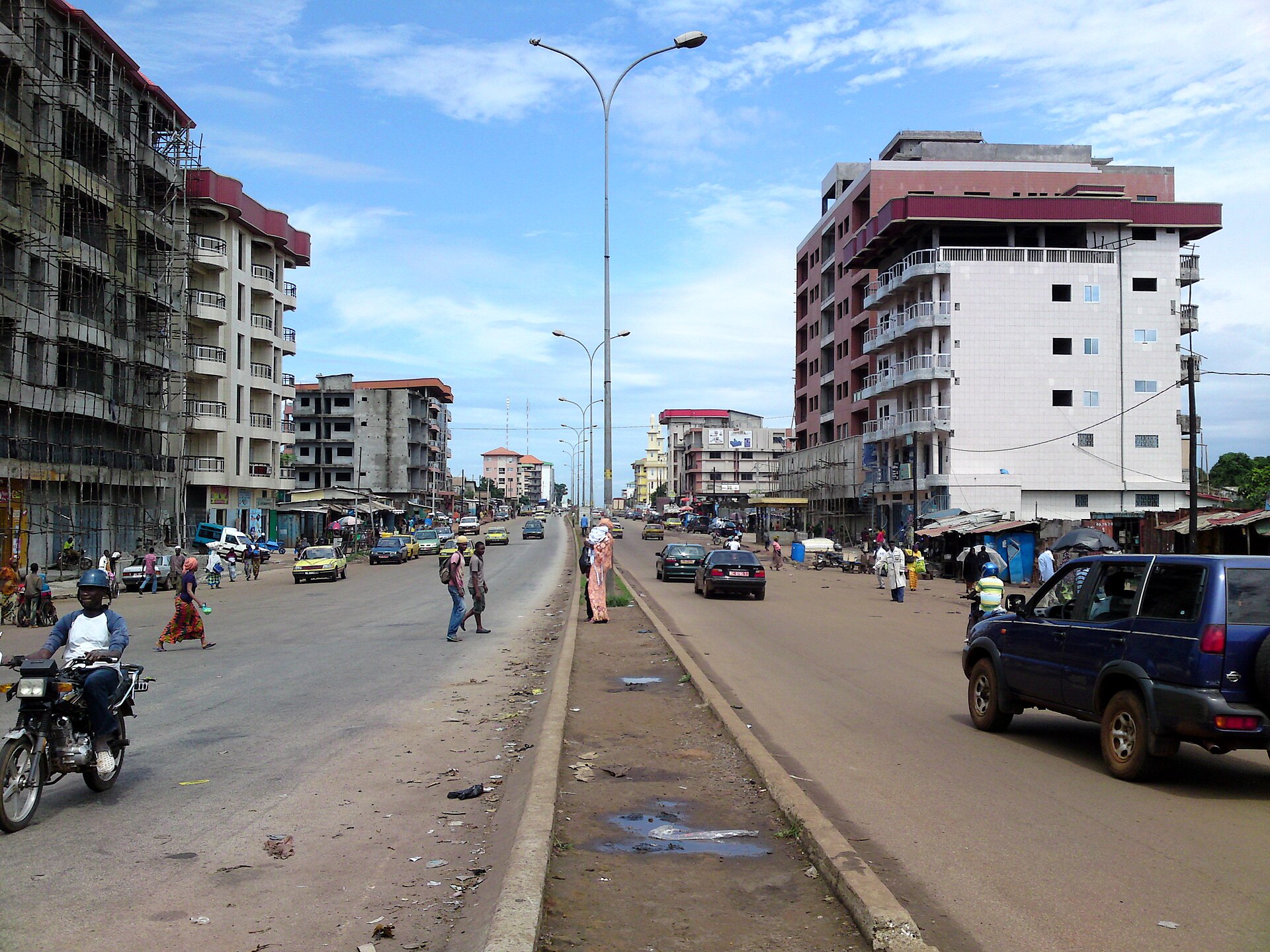 A busy avenue in Conakry with motorbikes, cars, pedestrians and apartment buildings under construction