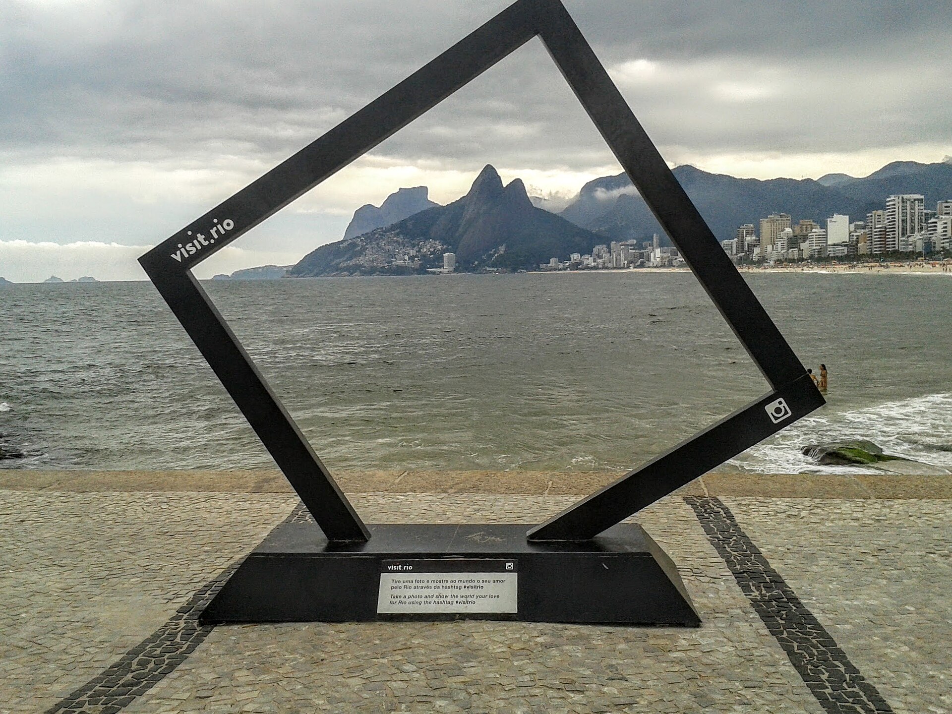 A black metal frame sculpture on the Arpoador waterfront in Rio de Janeiro, framing the Dois Irmaos mountain and the coast