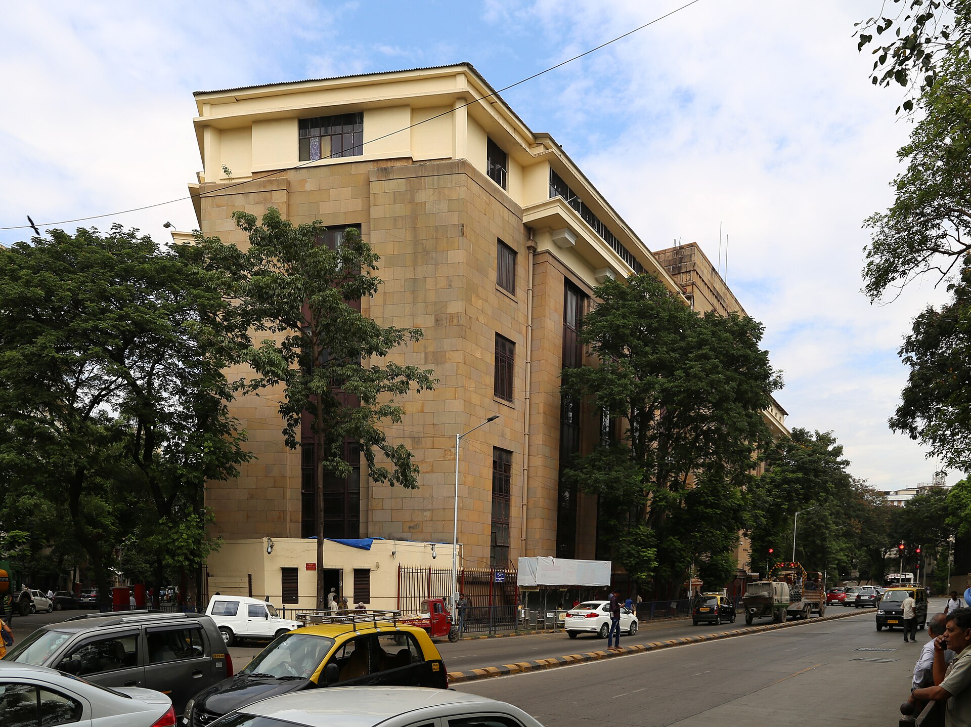 Side view of the Reserve Bank of India building on a Mumbai street with taxis and trees.