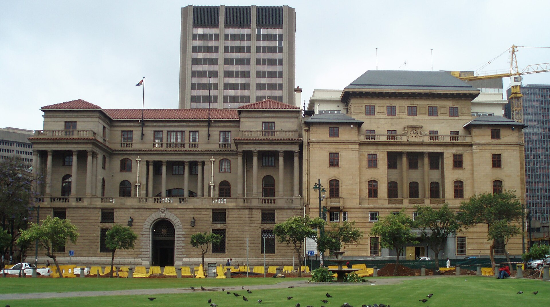 Two sandstone heritage buildings with columns and a red-tiled roof facing a lawn on Church Square, with a tall office tower behind