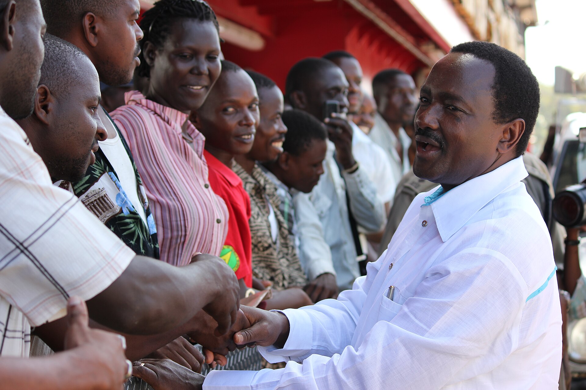 A man in a white shirt smiles and shakes hands with a line of supporters standing behind a railing in a street