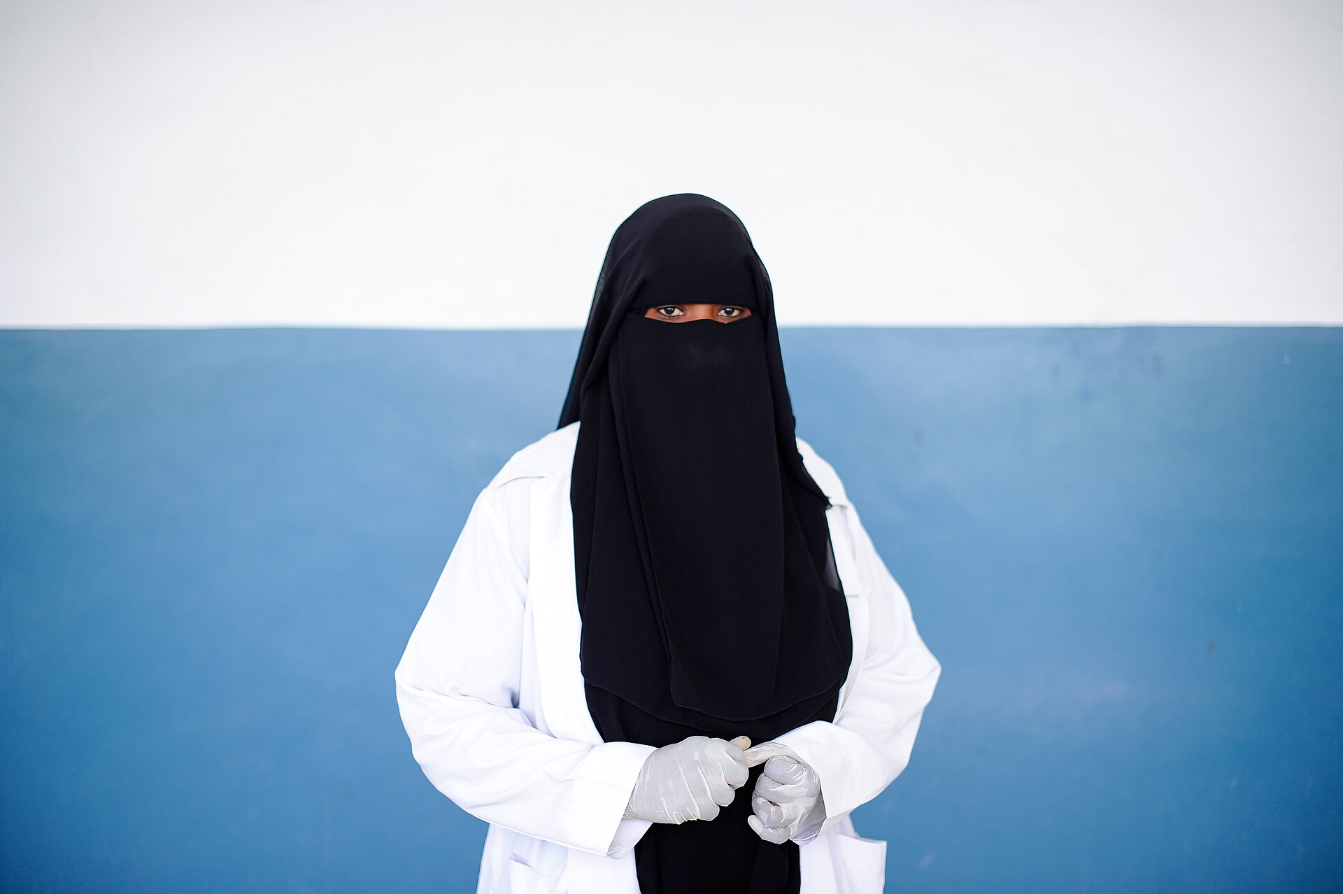 A nurse in a white coat, black niqab and latex gloves stands against a blue and white wall at Banadir hospital in Mogadishu